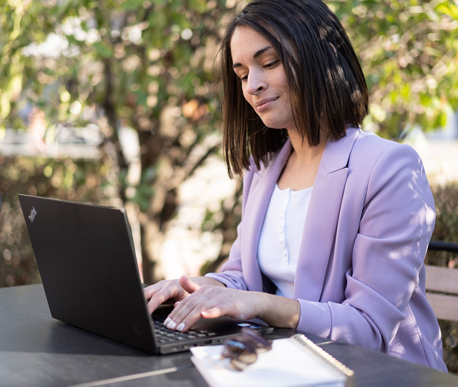 Frau mit Laptop sitzt an einem öffentlichen Platz in der Natur und arbeitet an einem Tisch am Laptop.